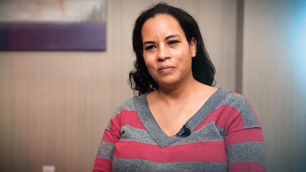 A woman with dark hair and a striped sweater sits indoors, looking at the camera with a slight smile.