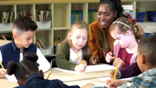 A teacher is assisting a group of young students writing at their desks in a classroom filled with bookshelves.