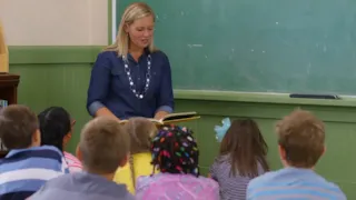 A teacher reads to a group of young students sitting on the floor in front of a chalkboard in a classroom.