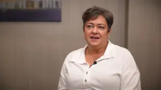 Person with short hair in a white shirt speaking, seated against a brown wall with a blurred painting in the background.