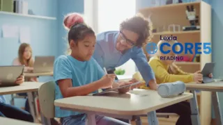 Teacher helps a student writing on a tablet in a classroom; other students work on laptops.