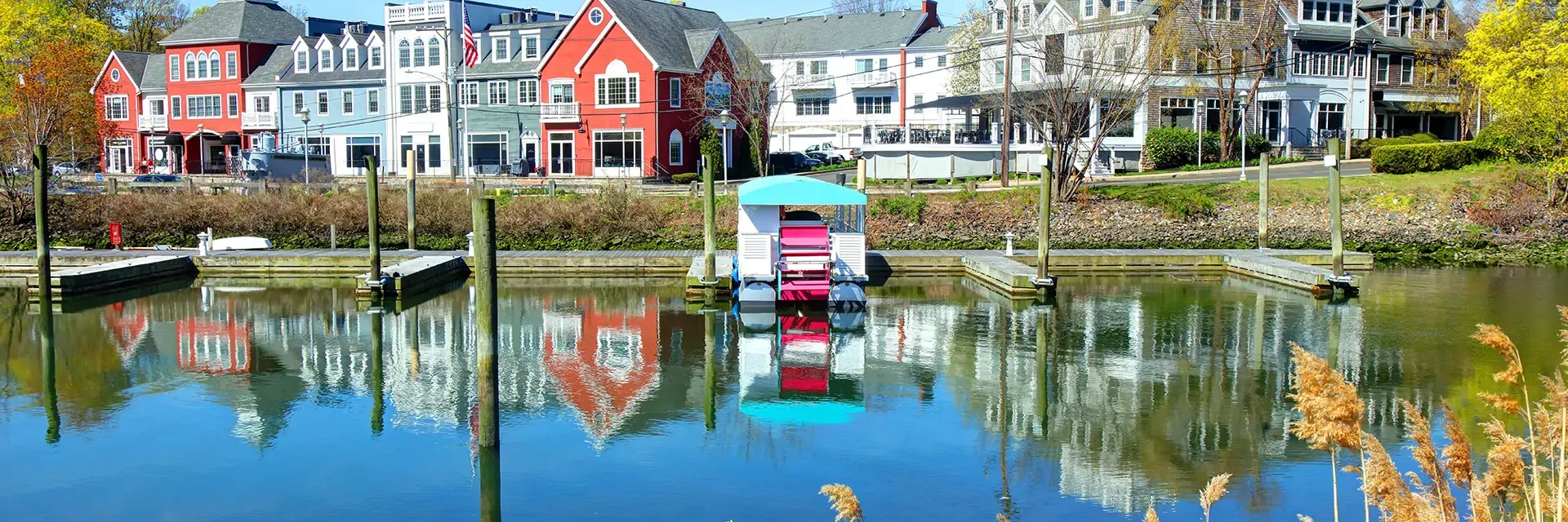 Colorful waterfront buildings reflect in a calm marina with empty docks and a small covered boat in the foreground.