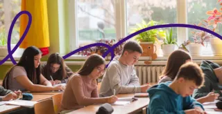 Students sitting at desks in a classroom, writing in notebooks, with sunlight and plants by the window.