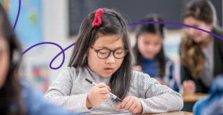 Young girl with glasses and red hair clip writing in a classroom, other students blurred in the background.