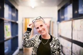 A person in a patterned jacket, smiling and holding glasses on their head, stands in a hallway lined with colorful artwork.