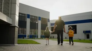 An adult walks hand in hand with two young children wearing backpacks toward a modern school building.