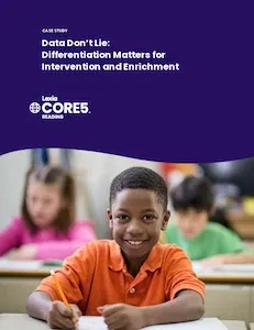 A smiling boy in an orange shirt writes at his desk in a classroom with two other children in the background.