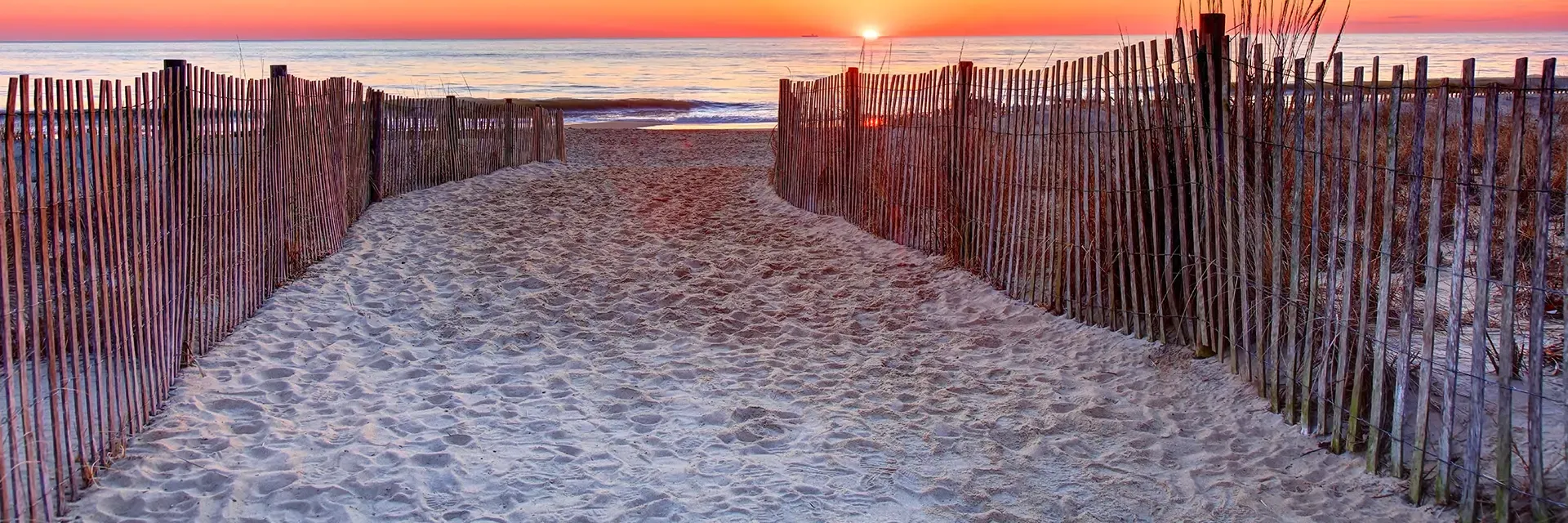 Sandy path between wooden fences leads to the beach at sunset, with the sun setting over the ocean horizon.