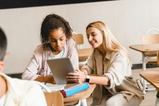 Teacher helping a student with a tablet in a classroom, both smiling. Desks and chairs are arranged around them.