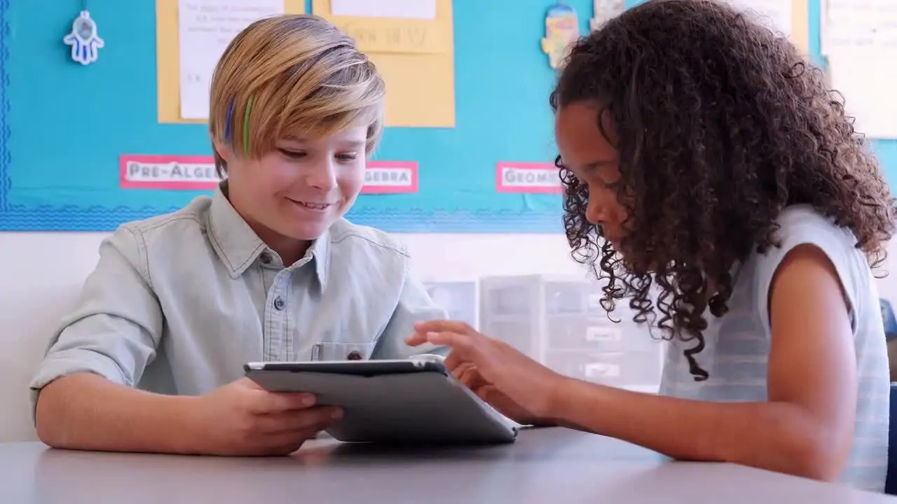 Two children sitting at a table, smiling and looking at a tablet together in a classroom setting.