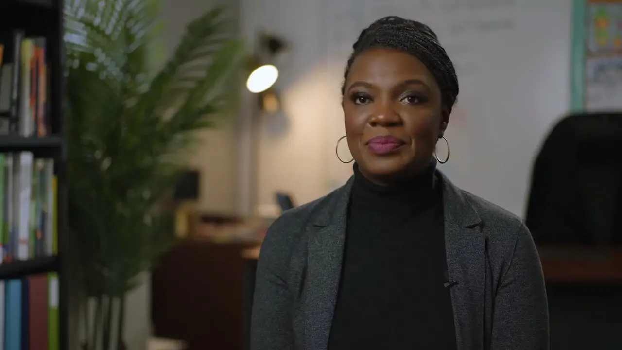 A woman in a gray blazer and black turtleneck smiles in an office with plants and bookshelves in the background.
