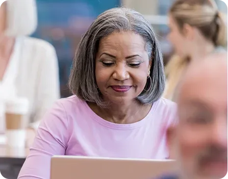 Older woman with gray hair and pink shirt using a laptop, seated in a busy indoor setting with others around her.