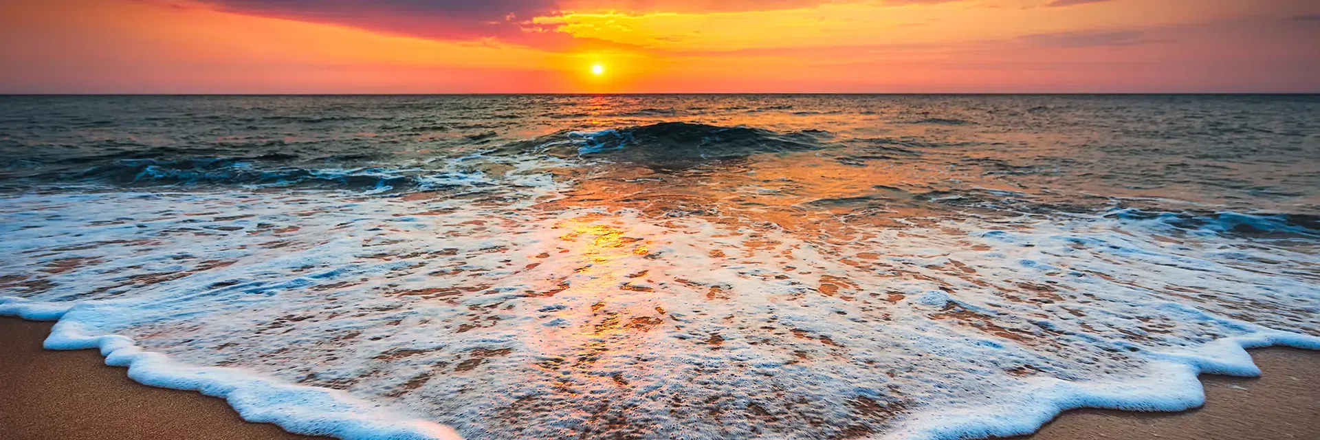 Sunset over the ocean with waves gently washing onto a sandy beach, sky glowing orange and pink.