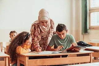 A teacher helps two students with their work at a desk in a bright classroom.