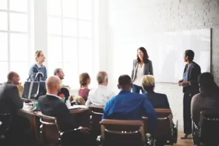 People in business attire attend a meeting; two women stand and present at a whiteboard in a bright office.