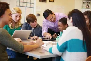 A teacher helps a group of diverse students working together around a table in a classroom setting.