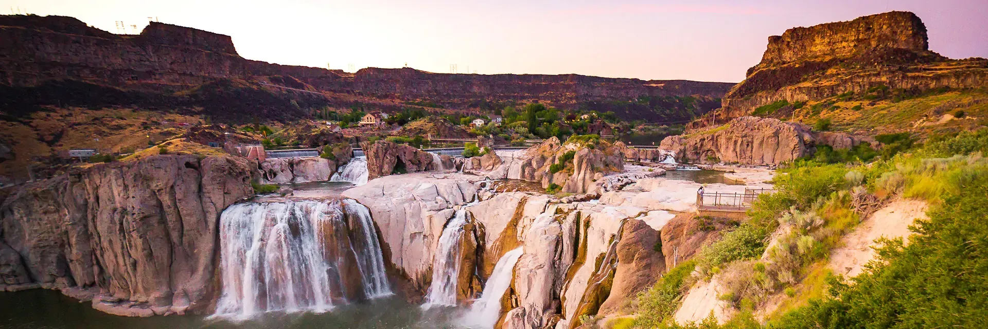 Wide view of Shoshone Falls cascading over rocky cliffs, surrounded by green vegetation and canyon walls at sunset.