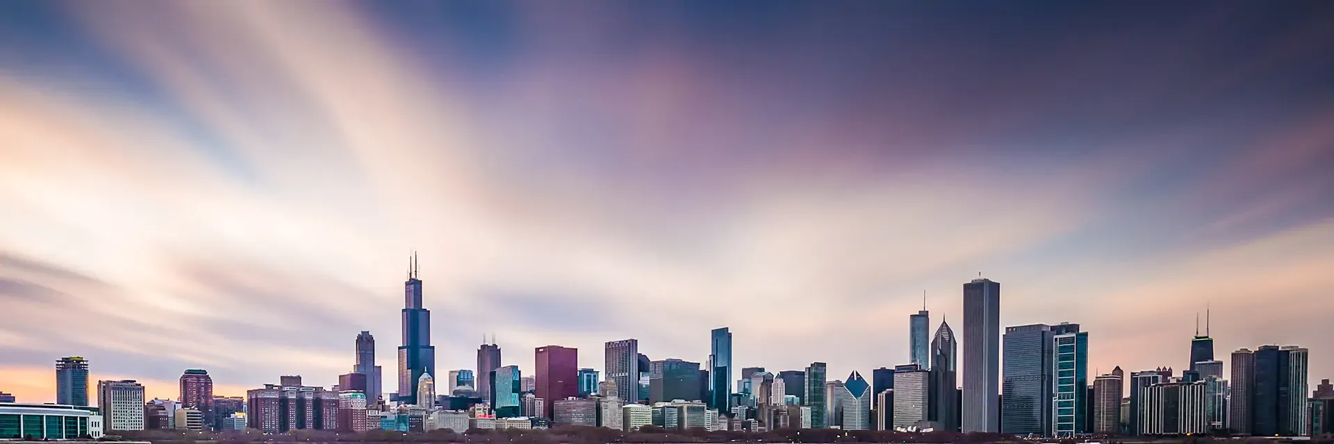 Chicago skyline at sunset with colorful clouds and tall buildings, including the Willis Tower and John Hancock Center.