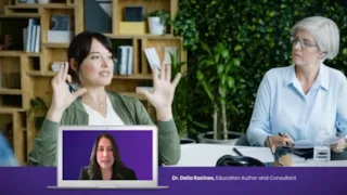 Two women sit at a table talking, with a laptop displaying a woman speaking in a video call on the screen.