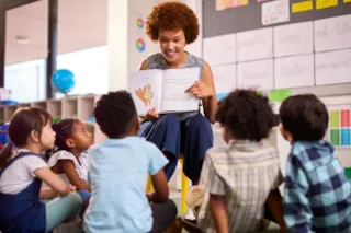 A teacher with curly hair reads a book to a group of young children sitting on the floor in a colorful classroom.