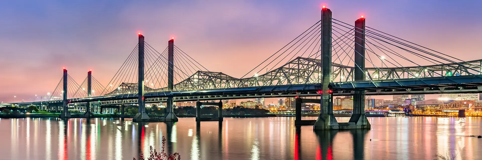 Cable-stayed bridge with illuminated towers at dusk, spanning a calm river with city lights in the background.