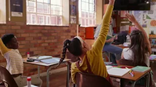 Students in a classroom sitting at desks with hands raised, attentively looking toward the front of the room.