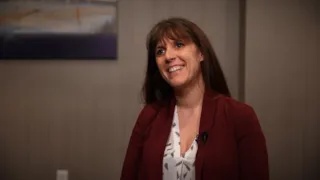 A woman with long brown hair smiles while sitting indoors, wearing a maroon blazer and white patterned blouse.