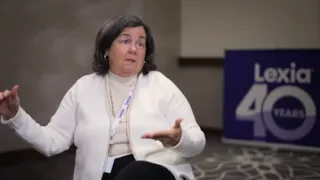 A woman in a white sweater speaks while seated indoors, with a Lexia 40 sign visible in the background.