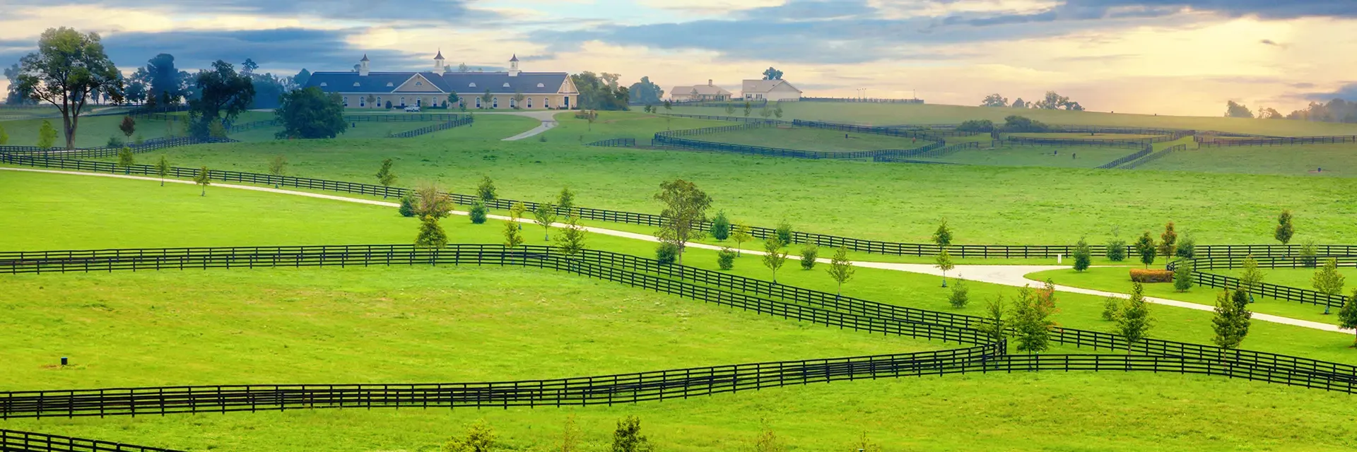 Green rolling hills with black wooden fences, a curving path, and a large barn under a partly cloudy sky.