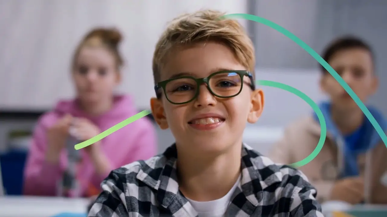 Smiling boy with glasses and plaid shirt sits in a classroom, with two students blurred in the background.