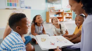 A teacher reads a picture book to a group of smiling and laughing children sitting on the classroom floor.