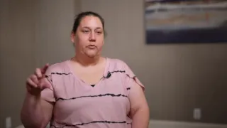 A woman in a pink shirt sits indoors, speaking, with a blurred painting on the wall behind her.