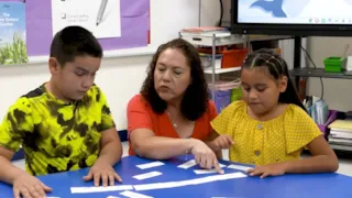 A teacher helps two children with a word sorting activity at a blue table in a classroom.
