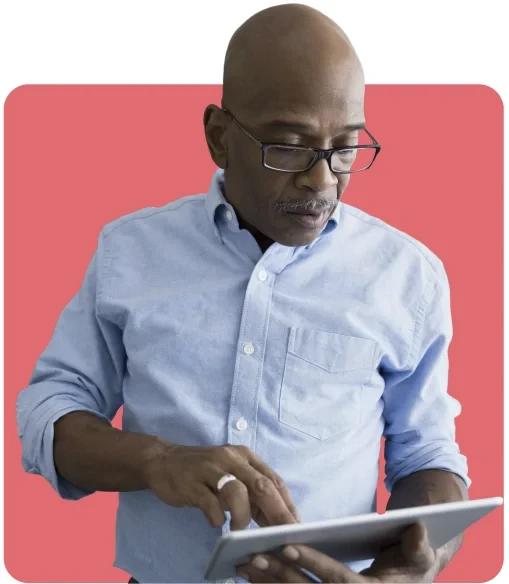 Man wearing glasses and a light blue shirt using a tablet against a solid pink background.