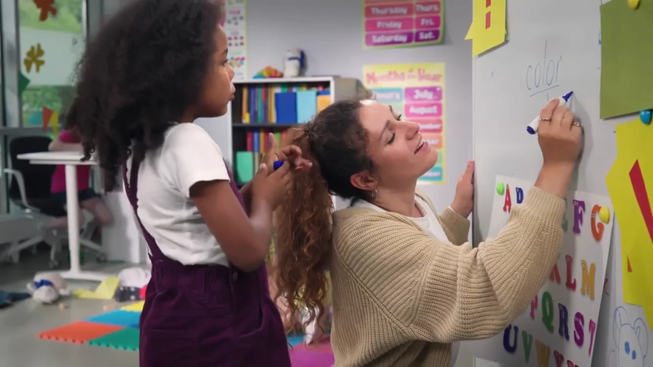 A young girl braids a teacher’s hair while the teacher draws on a whiteboard in a colorful classroom.