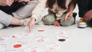 Three children sitting on the floor, pointing at scattered alphabet flashcards with pictures and words.