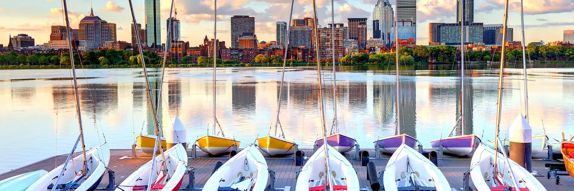 Sailboats lined up on a dock with a city skyline and water reflecting buildings at sunset.