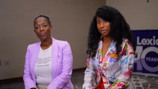Two women sit side by side, one in a lavender blazer, the other in a colorful printed blazer, facing the camera.