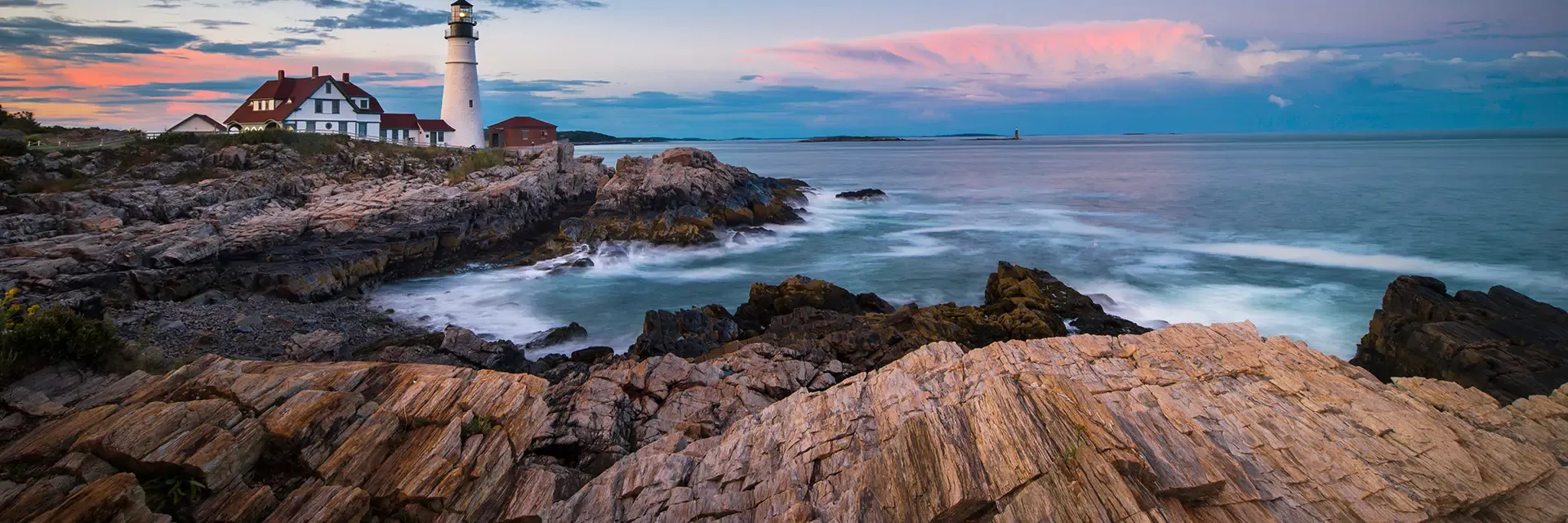 A lighthouse and house sit on rocky cliffs by the ocean at sunset, with waves crashing and clouds in the sky.