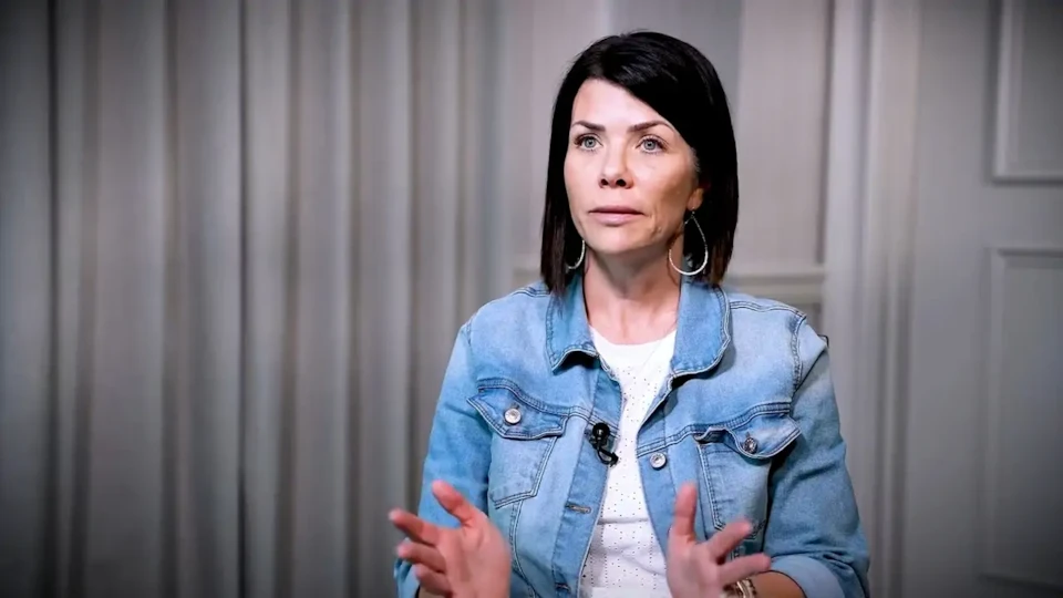 A woman with dark hair in a denim jacket gestures while speaking, seated indoors in front of light-colored curtains.