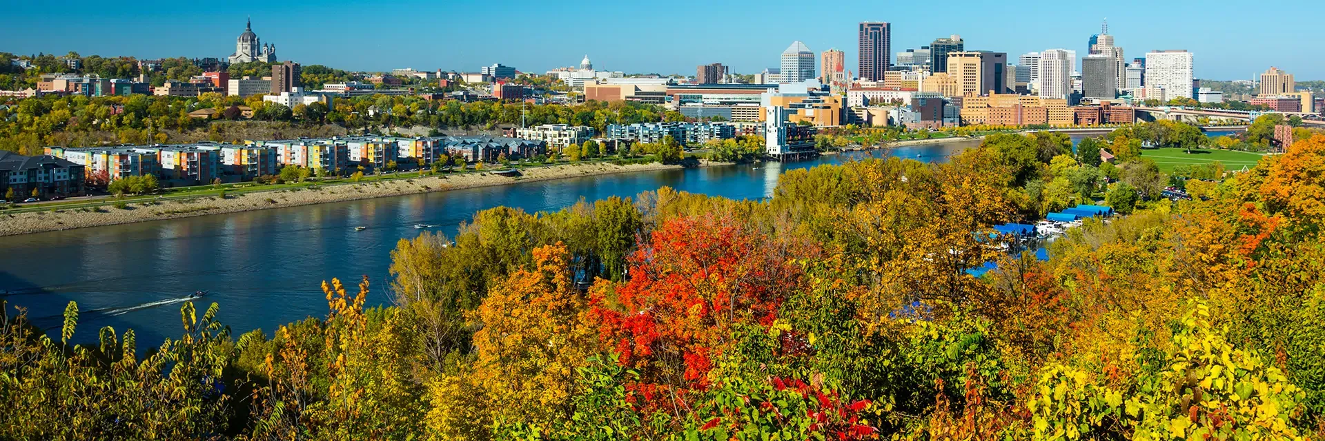 Panoramic view of a city skyline behind a river, with colorful autumn trees in the foreground.