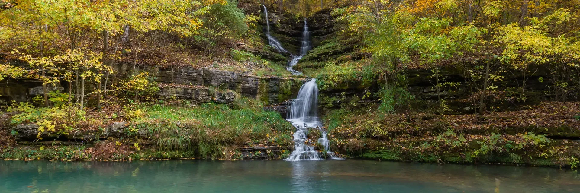 Small waterfall flows down rocky ledges into a calm pond, surrounded by lush green and yellow autumn trees.