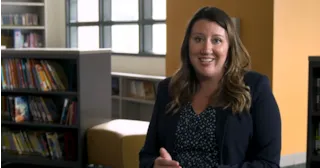 Woman with long brown hair sits smiling in a library, wearing a navy blazer and patterned blouse.