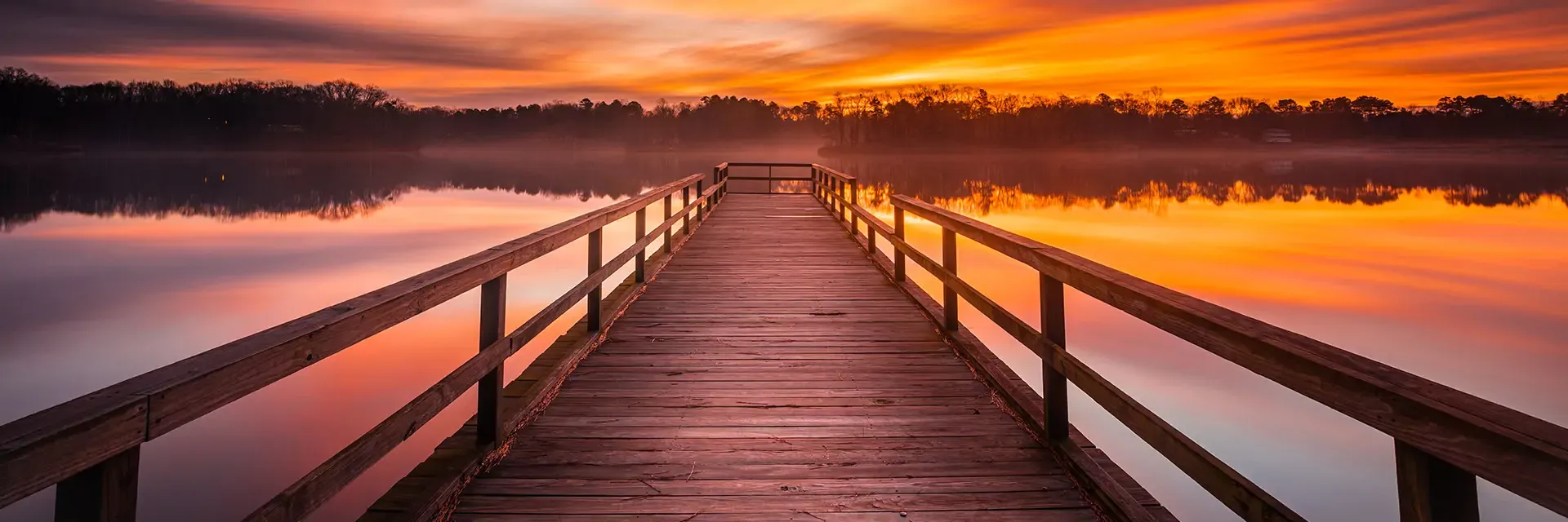 Wooden pier extends over calm lake with vibrant orange and purple sunrise reflecting in the water.