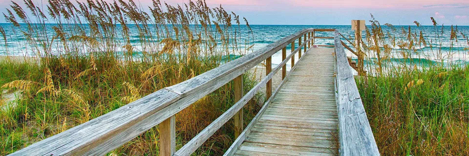 Wooden boardwalk leading through tall grass to a sandy beach with ocean waves under a pastel sky at sunset.