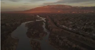 A winding river flows through a forest plain at sunset, with distant mountains under a cloudy sky.