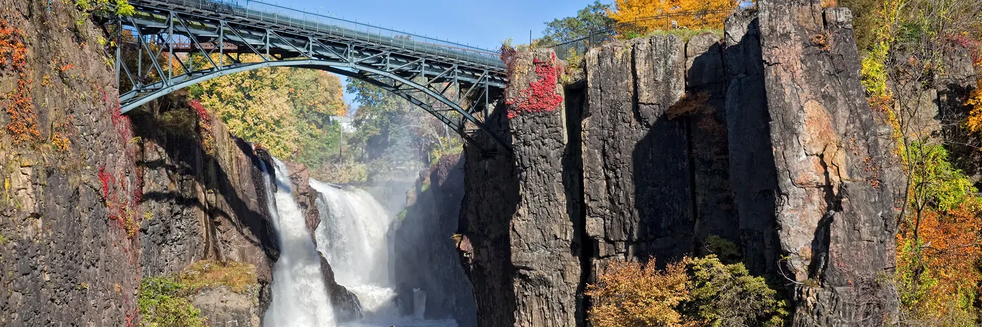 Waterfall flowing between tall rocky cliffs with a bridge above, surrounded by trees with autumn foliage.