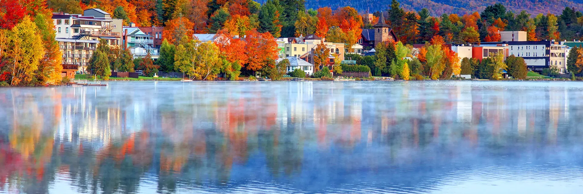 Colorful autumn trees and buildings reflected on a calm lake with mist rising from the water’s surface.