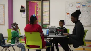 Three children and an adult sit at a table in a classroom, using laptops and headphones, engaged in learning activities.