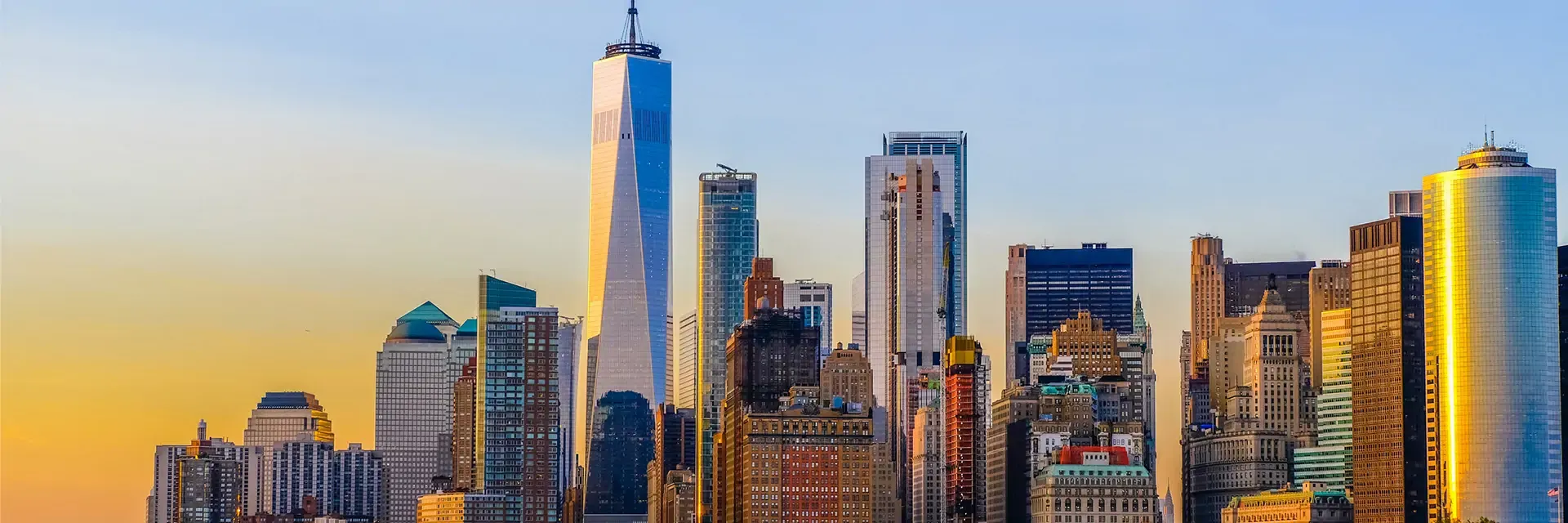 New York City skyline at sunset, featuring One World Trade Center and surrounding skyscrapers against a clear sky.